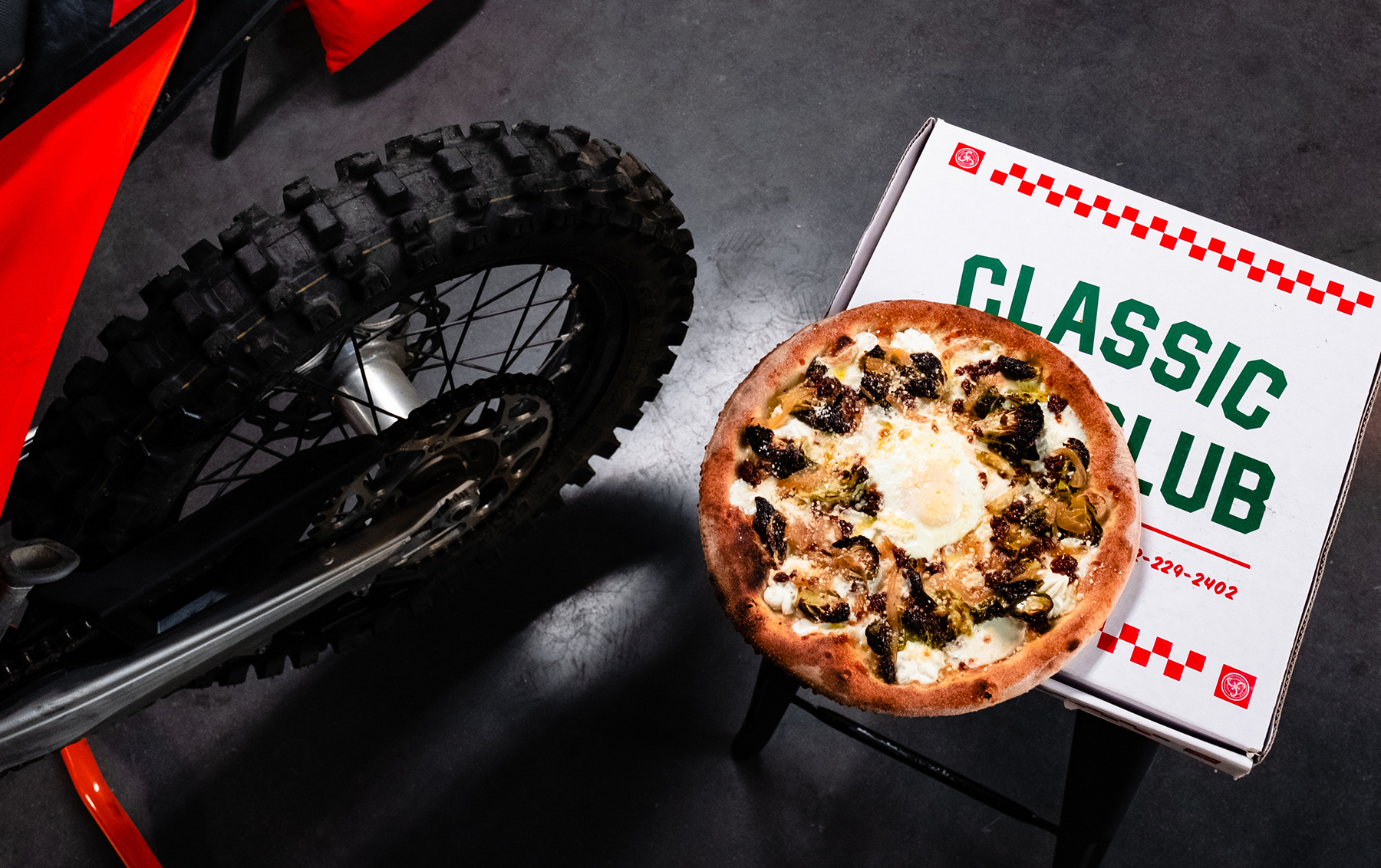 A lifestyle shot of a pizza sitting on top of the pizza box next to a dirtbike